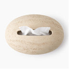 Light beige, pebble-shaped textured dispenser with an oblong opening from which white tissues emerge, isolated on white.
